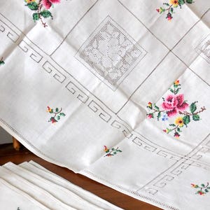 K&ouml;nnte beinhalten: Wei&szlig;es Leinentischtuch und passende Servietten, verziert mit gestickten rosa Rosen, gelben und blauen Blumen und gr&uuml;nen Bl&auml;ttern. Das Tischtuch hat ein geometrisches Muster und dekorative Lochmusterquadrate, ideal f&uuml;r eine Vintage-Tafel.