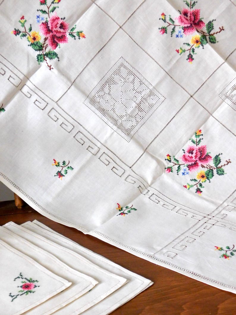 K&ouml;nnte beinhalten: Wei&szlig;es Leinentischtuch und passende Servietten, verziert mit gestickten rosa Rosen, gelben und blauen Blumen und gr&uuml;nen Bl&auml;ttern. Das Tischtuch hat ein geometrisches Muster und dekorative Lochmusterquadrate, ideal f&uuml;r eine Vintage-Tafel.