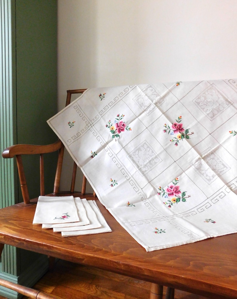 K&ouml;nnte beinhalten: Ein wei&szlig;es, besticktes Tischtuch und passende Servietten mit floralen Designs. Das Tischtuch hat ein Gittermuster mit gestickten Blumen in Rosa, Gelb und Blau. Die Servietten haben eine &auml;hnliche Blumenstickerei. Das Set wird auf einem Holztisch pr&auml;sentiert.