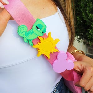 May include: A pink crossbody bag strap featuring three decorative silicone charms: a green chameleon, a yellow sun, and a light purple mouse-ear silhouette. The strap is worn over a white tank top and blue denim jeans, showcasing the colorful accessories.