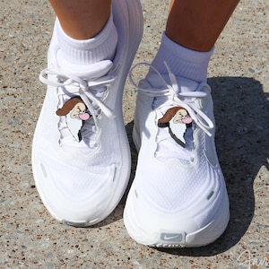 May include: A pair of white athletic sneakers featuring white socks and a small, colorful Grumpy character charm attached to the laces of each shoe. The sneakers have a mesh upper, white laces, and a small Nike logo on the front toe area.
