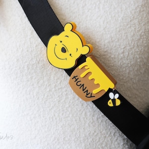 Puede incluir: Una cara amarilla de Winnie the Pooh, un tarro de miel con el texto "HUNNY" y un dije de abeja están unidos a una correa negra. La correa se usa sobre un suéter blanco.