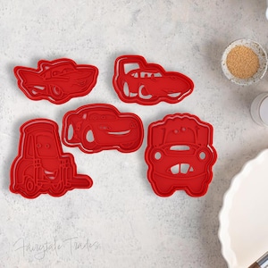 Può includere: Un set di cinque tagliabiscotti in plastica rossa raffiguranti vari personaggi a forma di auto con contorni dettagliati. I tagliabiscotti sono disposti su una superficie chiara accanto a utensili da forno, tra cui una frusta.