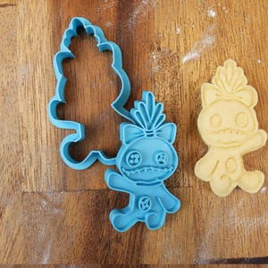 May include: A blue plastic cookie cutter and a baked cookie in the shape of a cartoon character. The cookie cutter is in the shape of the character's body and a separate tree-like shape. The baked cookie is light yellow.