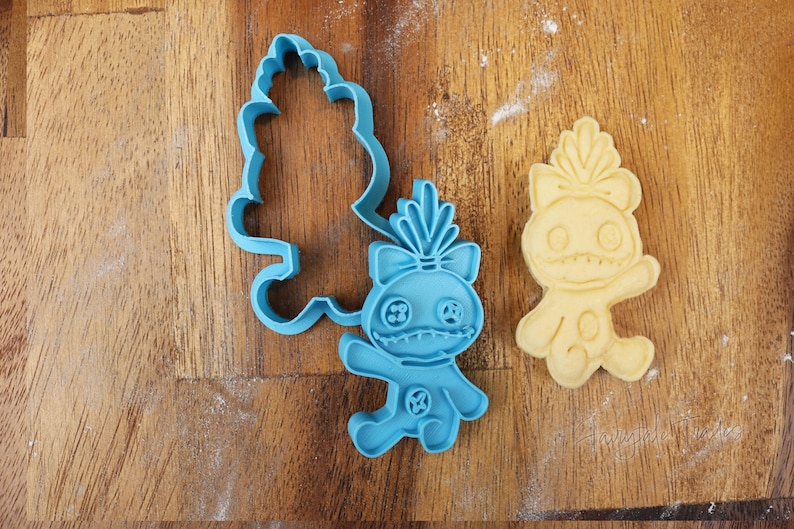 May include: A blue plastic cookie cutter and a baked cookie in the shape of a cartoon character. The cookie cutter is in the shape of the character's body and a separate tree-like shape. The baked cookie is light yellow.
