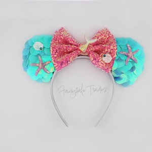 May include: A mermaid-themed headband with a silver band. The ears are covered in teal sequins, adorned with starfish and seashells. A large pink sequined bow sits in the center. The text "Fairytale Trades" is on the band.