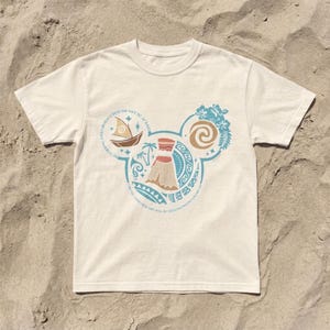 Op de afbeelding: Wit t-shirt met een cirkelvormig ontwerp met een vulkaan, een boot en bloemelementen in blauw, bruin en beige. Het ontwerp heeft de vorm van Mickey Mouse oren. Tekst rond het ontwerp luidt: "Wees moedig en wees niet bang."