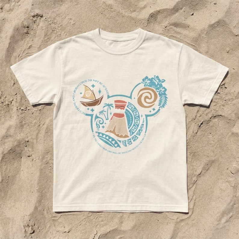 Op de afbeelding: Wit t-shirt met een cirkelvormig ontwerp met een vulkaan, een boot en bloemelementen in blauw, bruin en beige. Het ontwerp heeft de vorm van Mickey Mouse oren. Tekst rond het ontwerp luidt: "Wees moedig en wees niet bang."