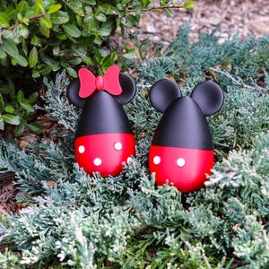 May include: Two decorative egg-shaped figures, one with a red bow, resembling Mickey and Minnie Mouse. The figures are black on top and red on the bottom with white polka dots. They are set among green foliage.
