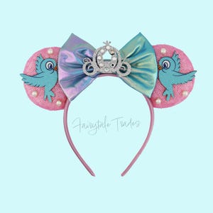 May include: Pink sequined mouse ears with a light blue bird applique and pearl accents. A holographic bow and a silver crown embellish the center. The headband is pink, and the text "Fairytale Trades" is visible.