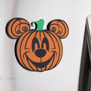 May include: A Halloween-themed magnet featuring a smiling pumpkin with Mickey Mouse ears. The pumpkin is orange with black details and a green stem. The magnet is attached to a stainless steel surface.