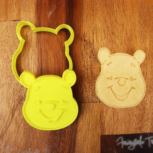 Op de afbeelding: Een gele Winnie de Poeh gezicht koekjesvorm naast een Winnie de Poeh koekje op een houten oppervlak. De koekjesvorm heeft de vorm van het gezicht van de beer met een lachende uitdrukking. De koek heeft hetzelfde lachende gezicht.