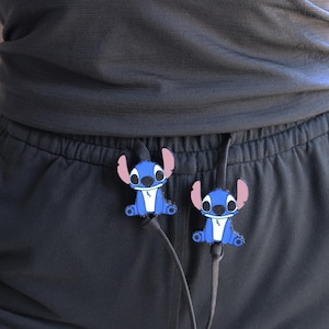 Op de afbeelding: Twee blauwe Stitch-vormige kabelorganizers bevestigd aan een zwart trekkoord. De stripfiguren hebben grote oren en zitten. De organizers zitten op een zwarte broek.