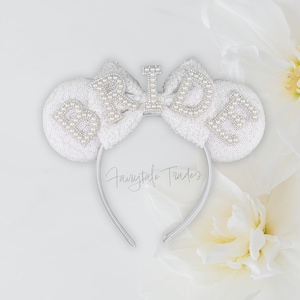 May include: White sequined headband with mouse ears, adorned with pearl-like beads spelling "Bride" and a central "I". The headband is silver-toned, with the text "Fairytale Trades" in a script font. The background features white flowers.