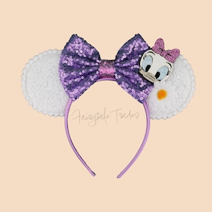 May include: A purple sequined bow headband with white sequined ears. The headband features a Daisy Duck character and a small pink bow. The text "Fairytale Trades" is visible on the headband.