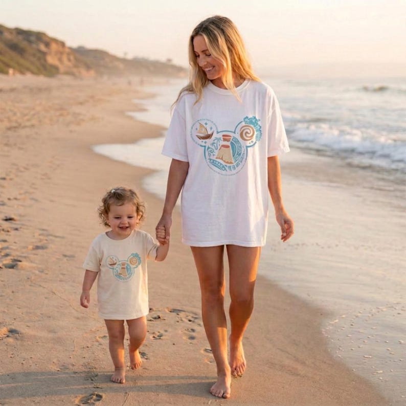 Op de afbeelding: Een vrouw en een kind lopen op een zandstrand. Beiden dragen T-shirts met een Mickey Mouse-vormig ontwerp met een boot, vuurtoren en werveling. Het shirt van de vrouw is wit, dat van het kind beige.
