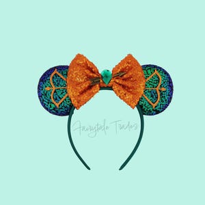 Op de afbeelding: Een teal hoofdband met pailletten muisoren en een oranje pailletten strik. De oren zijn versierd met een teal en goud design. Een groene edelsteen is gecentreerd op de strik. De tekst "Fairytale Trades" staat op de hoofdband.