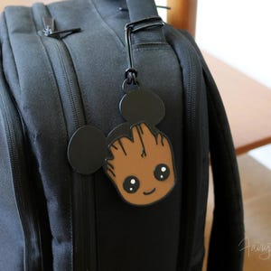Könnte beinhalten: Schwarzer Rucksack mit einem Gepäckanhänger im Groot-Design. Der Anhänger zeigt ein braunes Groot-Design mit einem lächelnden Gesicht und schwarzen Mickey Mouse-Ohren. Der Anhänger ist am Griff des Rucksacks befestigt.