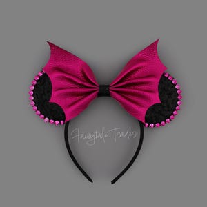 Puede incluir: Una diadema con dos orejas circulares negras con bordes tachonados de color rosa y un gran lazo texturizado de color rosa intenso con forma de alas de murciélago. El centro del lazo es negro y el texto Fairytale Trades está escrito en blanco.