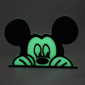 May include: A black and green glow-in-the-dark Mickey Mouse silhouette peeking over a surface. The silhouette is outlined in black and the inside is filled with a bright green glow.