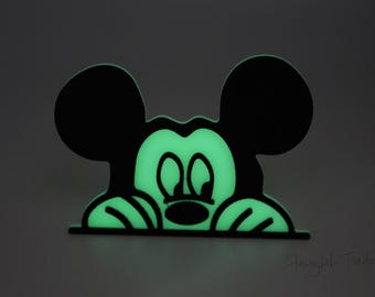 Op de afbeelding: Een zwart en groen, in het donker lichtend Mickey Mouse-silhouet dat over een oppervlakje gluurt. Het silhouet is zwart omlijnd en de binnenkant is gevuld met een heldergroene gloed.
