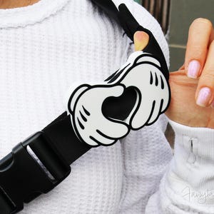 May include: A black and white Mickey Mouse hand-shaped accessory attached to a black strap with a buckle. The accessory is white with black outlines and details. The strap is worn over a white textured sweater.