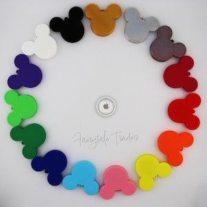 May include: A colorful arrangement of Mickey Mouse head-shaped accessories in various colors, including white, black, gold, silver, brown, red, orange, yellow, pink, blue, green, and navy. An Apple AirTag is in the center.