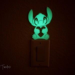 Könnte beinhalten: Ein leuchtendes grünes Stitch-Nachtlicht, geformt wie die Figur aus dem Zeichentrickfilm, ist über einem Lichtschalter angebracht. Das Licht strahlt in einem dunklen Raum sanft. Das Produkt ist ein dekorativer Artikel.