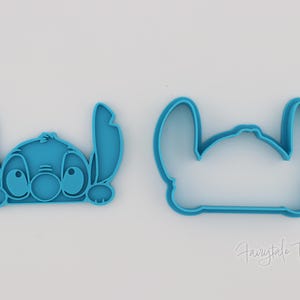 May include: Two blue plastic cookie cutters in the shape of the Disney character Stitch. One cutter is a detailed version of Stitch's face, and the other is an outline of the character's head. The cutters are on a white background.