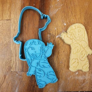 May include: A blue plastic cookie cutter in the shape of a cartoon character, next to a cookie cut in the same shape. The cookie cutter is on a wooden surface. The cookie is light yellow.