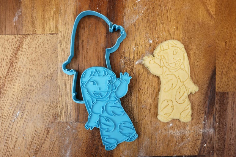 May include: A blue plastic cookie cutter in the shape of a cartoon character, next to a cookie cut in the same shape. The cookie cutter is on a wooden surface. The cookie is light yellow.
