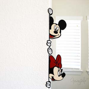 May include: A wall decal featuring Mickey Mouse and Minnie Mouse peeking around a corner. Mickey has a red mouth and Minnie has a red bow. The decal is black, white, and red.