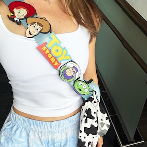 Könnte beinhalten: Eine hellblaue Schärpe mit dem Toy Story-Logo und Charakter-Icons von Jessie, Woody, Buzz Lightyear und einem Alien. Die Schärpe hat unten einen Akzent aus schwarz-weißem Kuhfellmuster-Stoff und wird diagonal über einem weißen Tanktop getragen.