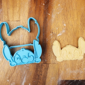 May include: A light blue plastic cookie cutter in the shape of the Disney character Stitch, next to a baked cookie in the same shape. The cookie cutter is on a wooden surface.