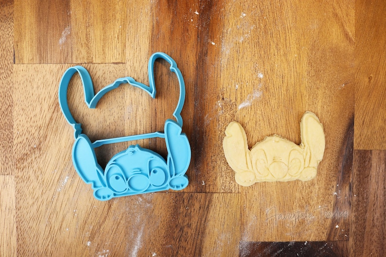May include: A light blue plastic cookie cutter in the shape of the Disney character Stitch, next to a baked cookie in the same shape. The cookie cutter is on a wooden surface.
