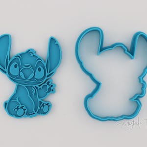 May include: Two teal-colored cookie cutters in the shape of the Disney character Stitch. One cutter is a solid figure of Stitch, and the other is an outline. The cutters are on a white background.