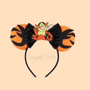 Peut inclure: Un serre-tête noir avec des oreilles rayées orange et noires. Un nœud noir à paillettes est placé entre les oreilles, avec le personnage de Tigrou au centre. Le texte "Fairytale Trades" est écrit sur le serre-tête.