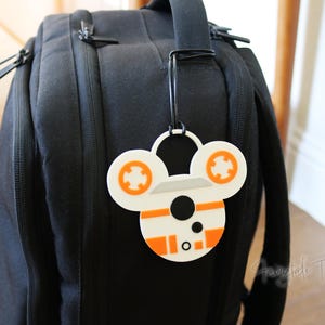 May include: A white and orange character-themed luggage tag is attached to a black backpack. The tag features a circular design with orange and white details. The backpack has multiple zippers and a handle.