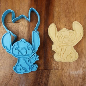 May include: A blue plastic cookie cutter in the shape of the character Stitch from the Disney movie Lilo & Stitch, next to a baked cookie in the same shape. The cookie cutter is on a wooden surface.