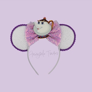 Può includere: Una fascia stravagante con un personaggio di teiera dei cartoni animati, un fiocco di paillettes e orecchie bianche con finiture viola. La fascia è argentata e lo sfondo è lilla chiaro.
