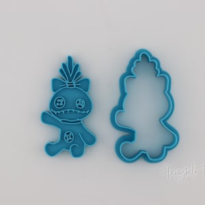 May include: Two blue plastic cookie cutters. One is a detailed figure of a cartoon character with a bow, and the other is the outline of the same character. The cutters are on a white surface.
