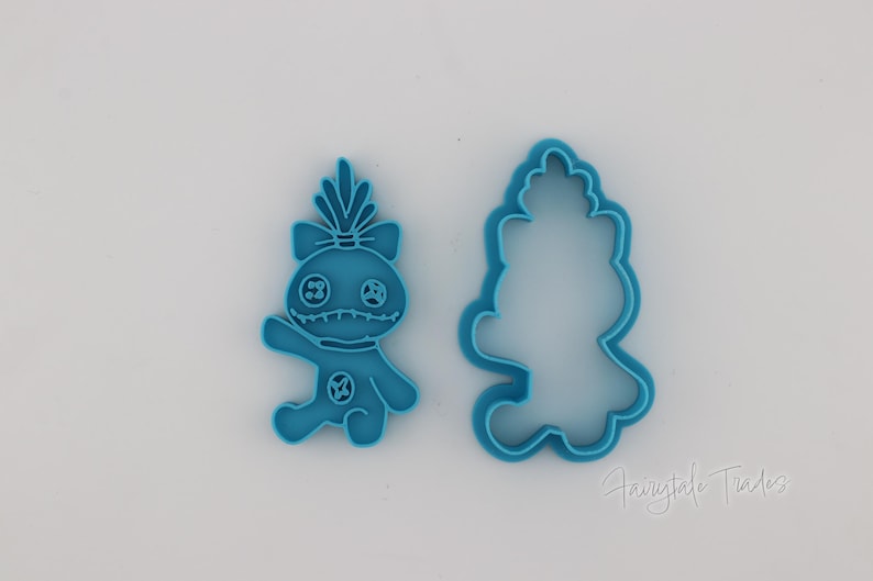 May include: Two blue plastic cookie cutters. One is a detailed figure of a cartoon character with a bow, and the other is the outline of the same character. The cutters are on a white surface.
