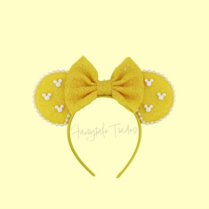 May include: Yellow mouse ear headband with a large bow and white Mickey Mouse accents. The ears are outlined with white pearls. The headband is yellow and the text "Fairytale Trades" is visible.