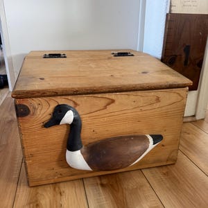 May include: A rectangular wooden storage box with a hinged lid and a prominent three-dimensional Canada goose figure attached to the front. The box features a natural light brown wood grain finish, black metal hinges, and a painted goose.