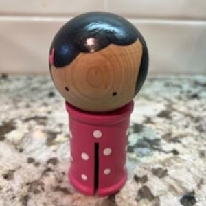 May include: Wooden doll with a round head and a pink polka-dotted base. The doll's head is painted with black hair and a light skin tone. The base is a cylinder shape with white polka dots and a vertical slit.
