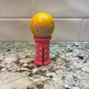 May include: A wooden doll with a yellow head and a pink body. The head is round with a painted face and yellow hair. The pink body is cylindrical with white polka dots.