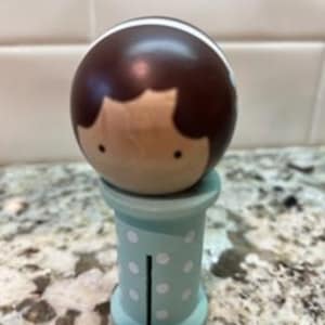 May include: A wooden peg doll featuring a round head with dark brown hair and a white headband, resting on a light blue cylindrical base decorated with small white polka dots and a vertical black line detail on the front of the base.