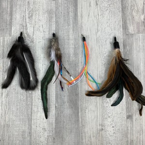 May include: Four cat toys with feathers in various colors. The toys have a black top with a metal loop for hanging. The feathers are black, gray, brown, green, and multicolored. The toys are arranged on a light gray wooden surface.
