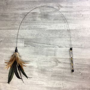 May include: A black and silver cat toy with a long, flexible wire and a feather attachment. The feather is brown, black, and tan.