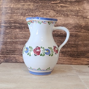 May include: A white ceramic pitcher with a blue and gold rim and floral designs in red, blue, and green.
